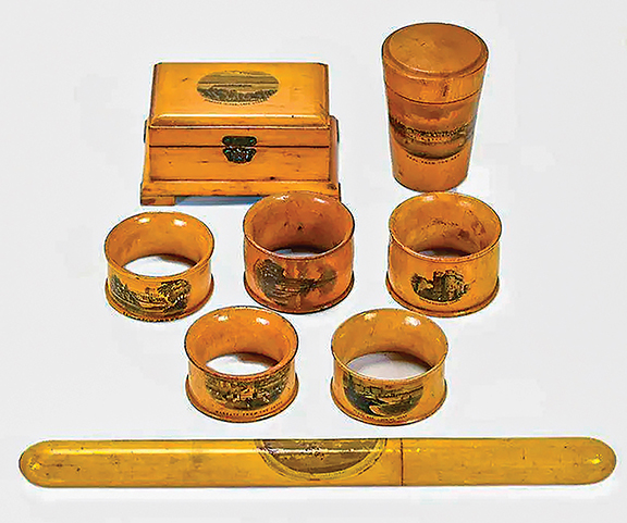 This lot consisted of eight pieces of late 19th-century Mauchline ware. There are five napkin rings, a needle case, and two boxes—one a tall cylindrical box and the other rectangular. Each of the pieces displays a transfer-printed scene. The scenes are locations and attractions in the United Kingdom, with the exception of the rectangular box, which depicts Diamond Island, Lake George, New York. The 9½