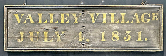 This double-sided wood sign is lettered “Valley Village / July 4, 1831.” It is painted black and yellow, with a molded frame, 12