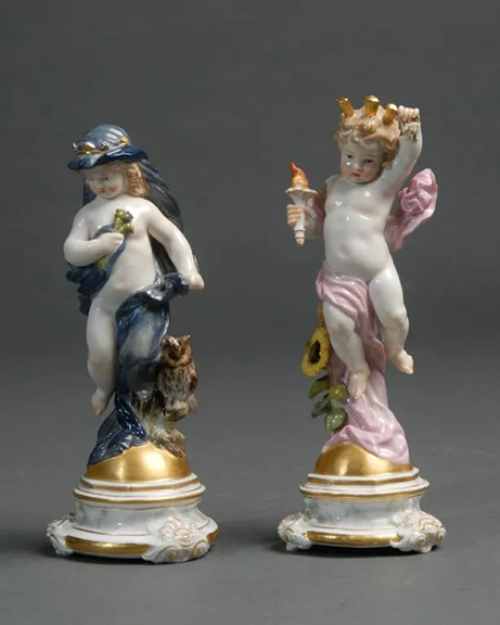 One of these Meissen porcelain putti represents day, and the other represents night. Day is positioned beside a large sunflower, draped in pink and holding a torch in one hand and a rose in the other. Night is modeled with an owl at his side, draped in blue, wearing a crown and holding a small bouquet of flowers. Each of the approximately 7