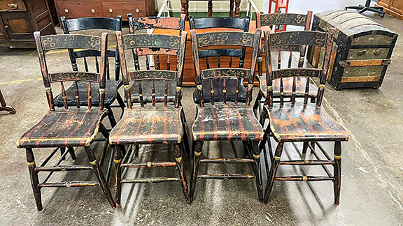 Three of these eight plank-seat thumb-back farmhouse chairs have been painted black. Five retain at least a portion of their original decorative scheme: black and red paint with gold or yellow accents. The medial back splats display a stenciled floral design. The top rails appears to show a combination of free and stenciled designs. The chairs appear to be structurally sound. The set sold for only $36.73.