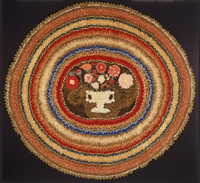 A Circular Shirred Rug With a Stylized Urn of Flowers  American, found in New England, circa 1840. Wool stitched on to linen.  A small urn filled with colorful flowers is used as a focal point of a vibrant aura of multi-colored concentric circles.  This rug represents a rare example of the shirring technique used within a circular format and is truly an extraordinary example of rug-making and folk art. 43½ in. x 41 in.  Illustrated: “American Hooked and Sewn Rugs: Folk Art Underfoot” by Joel & Kate Kopp. pg. 33, fig. 32. “The Needle Arts” Time Life Books, A Social History of American Needlework, pg. 116. Provenance: Stephen Score, Virginia Ramsey-Pope Cave, Northeast Auctions- August 5, 2000.
